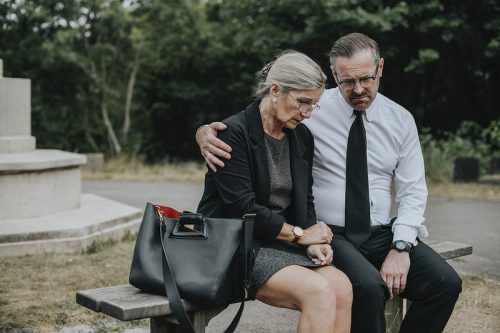 Elderly couple sitting on a park bench, with the husband reassuring his wife amidst a somber atmosphere.